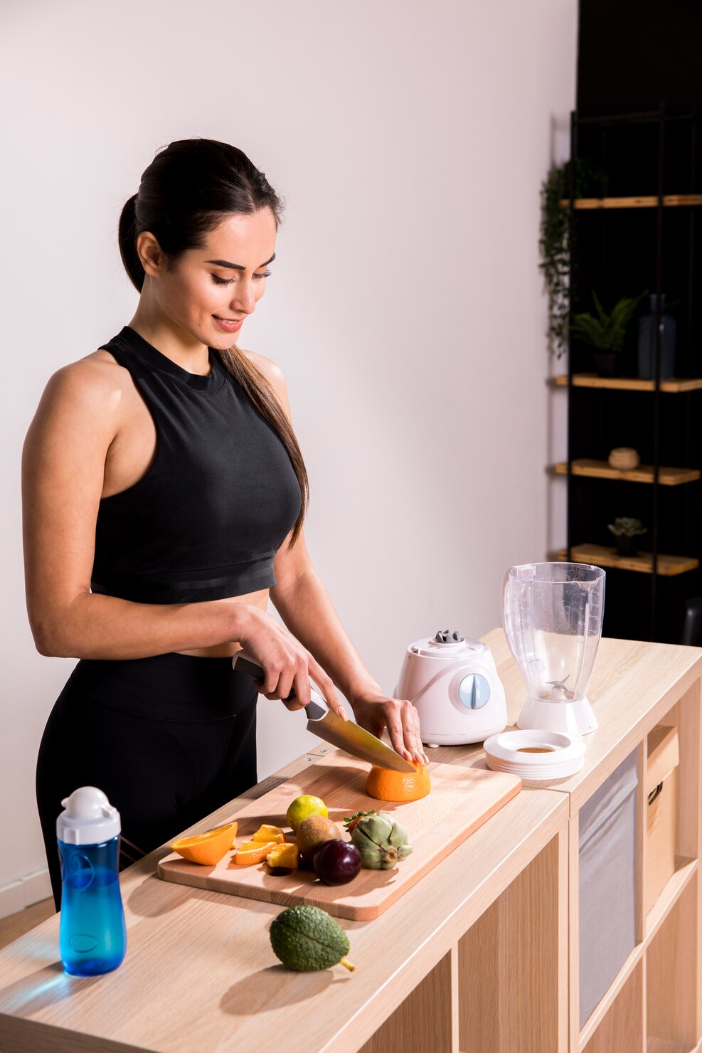 A woman in black workout clothes slices fruit on a kitchen counter beside a blender and a bottle of water.