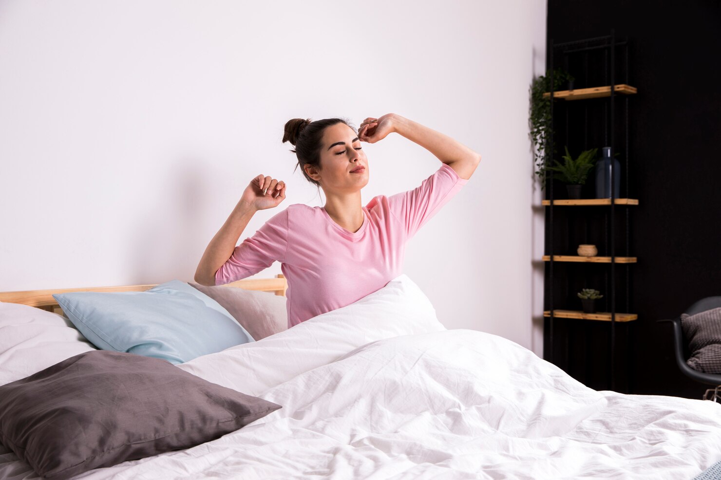 A woman sits up in bed in the morning and stretches both arms, surrounded by white bedding and pillows.