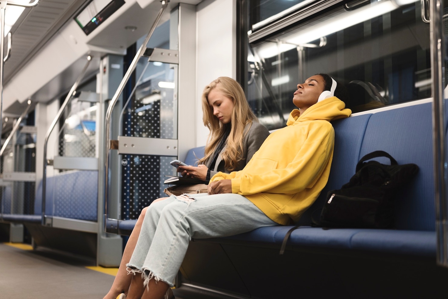 A subway passenger in a yellow hoodie sleeps beside another passenger using her phone.