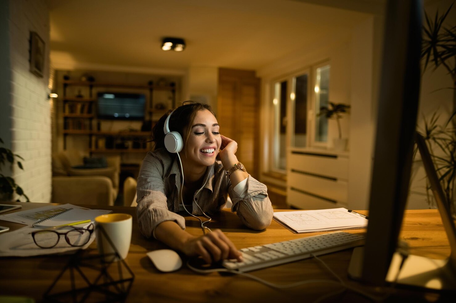 A woman wearing headphones smiles during a video call at a computer in a warmly lit home workspace.