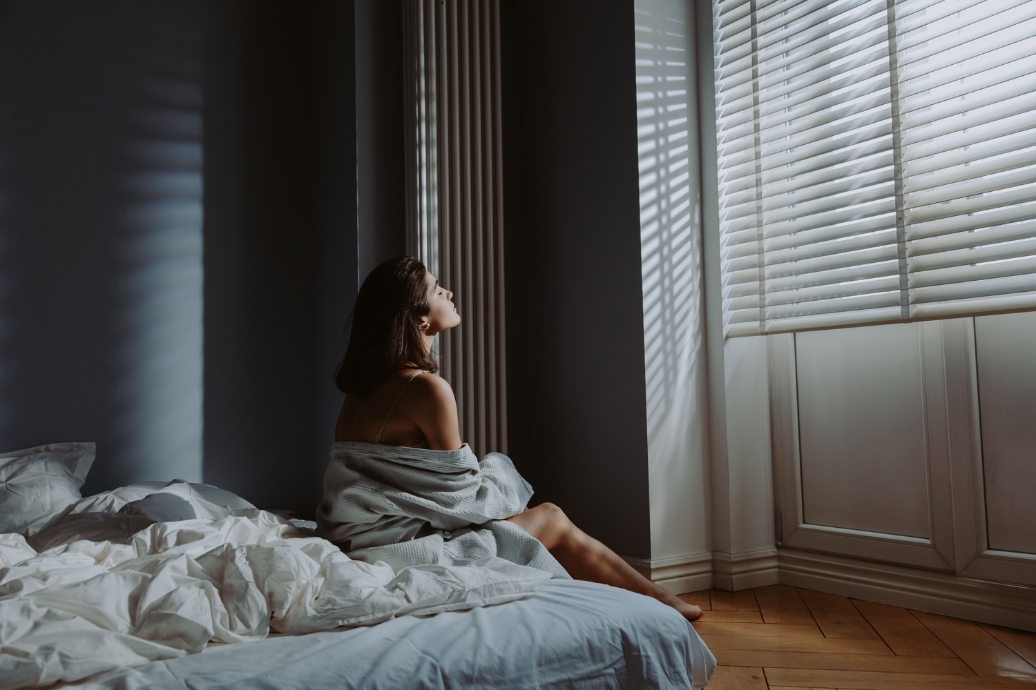 A woman wrapped in a blanket sits on an unmade bed as strong daylight cuts through the window blinds.