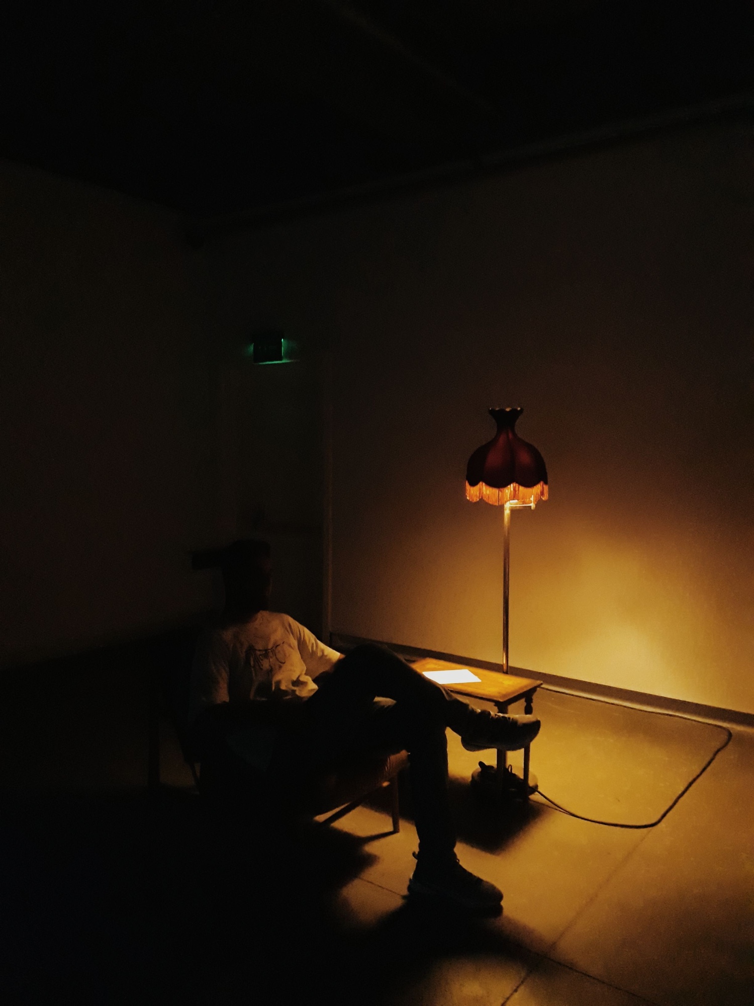 A person sits quietly in a dark room beside a single low lamp casting warm light and long shadows.