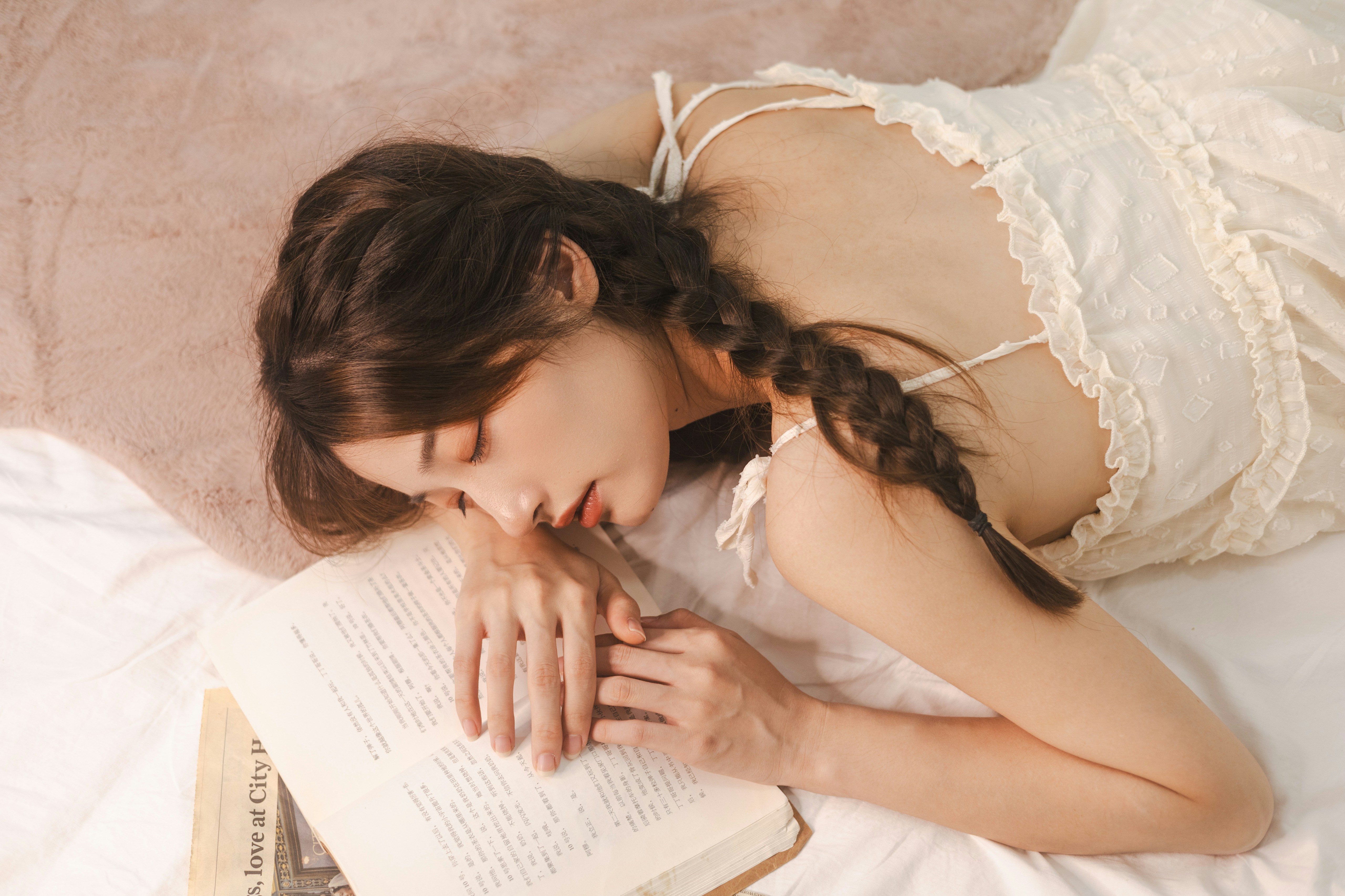 A young woman with long brown hair in two braids lies on her stomach on a bed, wearing a white sleeveless dress with ruffled edges. She is reading an open book, her hands resting gently on the pages. The bed is covered with white sheets and a beige blanket, and the environment feels soft, peaceful, and relaxed, suggesting a quiet moment of rest or study. Visible text on the book includes the phrase love at City.