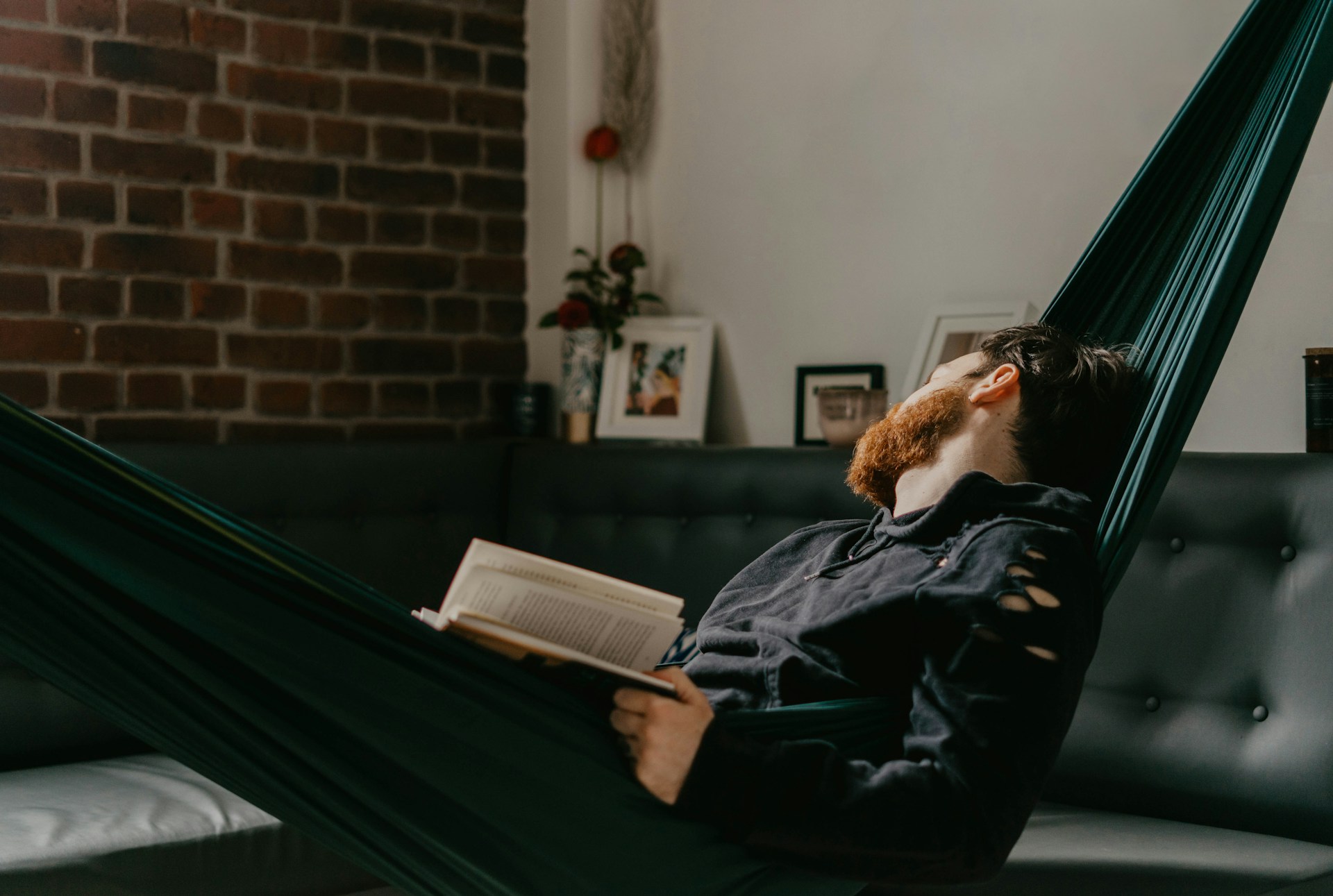 A man with a beard relaxes in a dark green hammock indoors, holding an open book in one hand. He gazes upward, partially reclined, wearing a black sweatshirt with torn sleeves. The room features a gray tufted sofa, exposed brick wall, framed photos, and a vase with flowers, creating a cozy and tranquil atmosphere.
