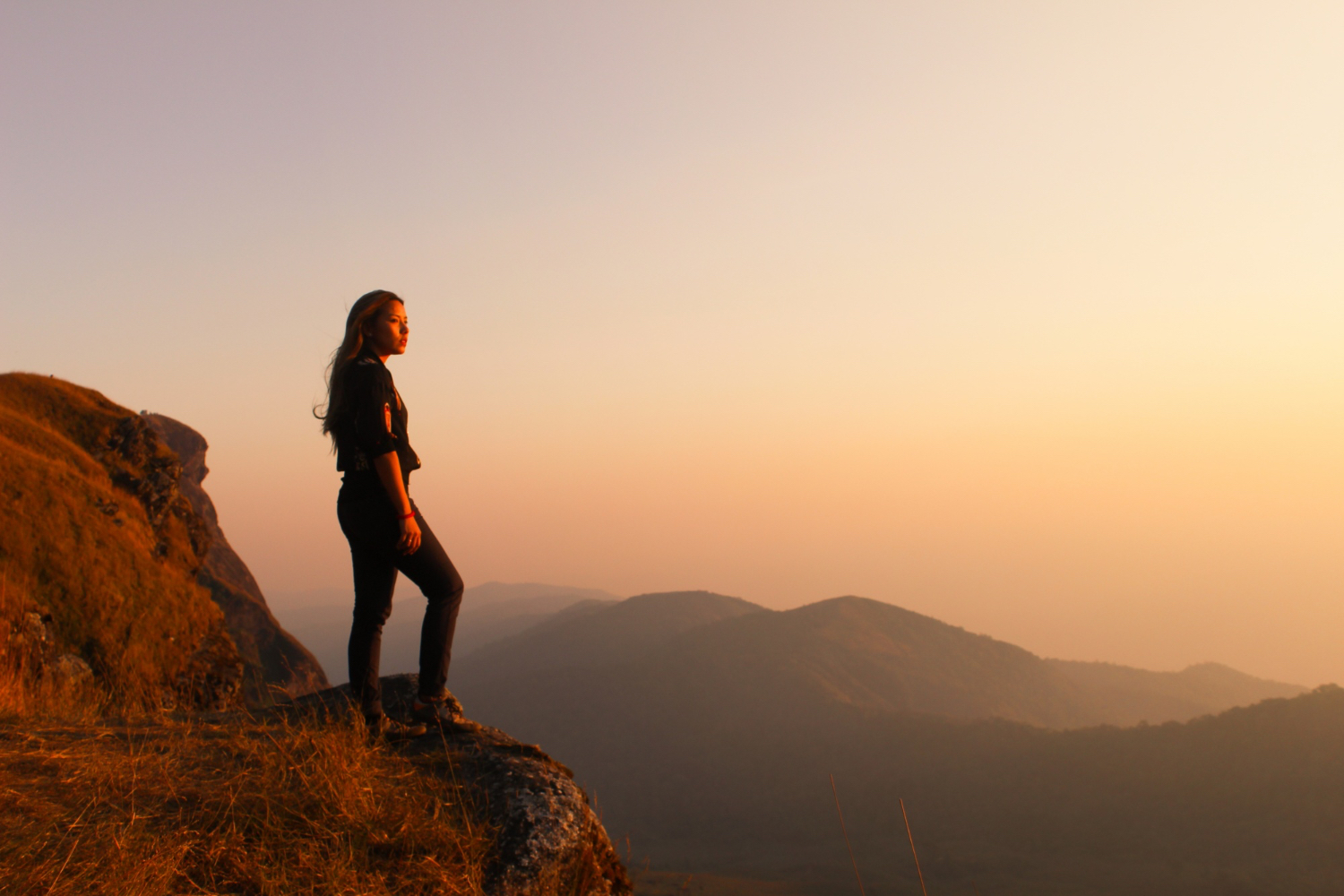 Person standing on the edge of a grassy cliff at sunset, gazing out over rolling hills and distant mountains. The sky is filled with warm orange and pink hues, creating a peaceful and contemplative atmosphere.