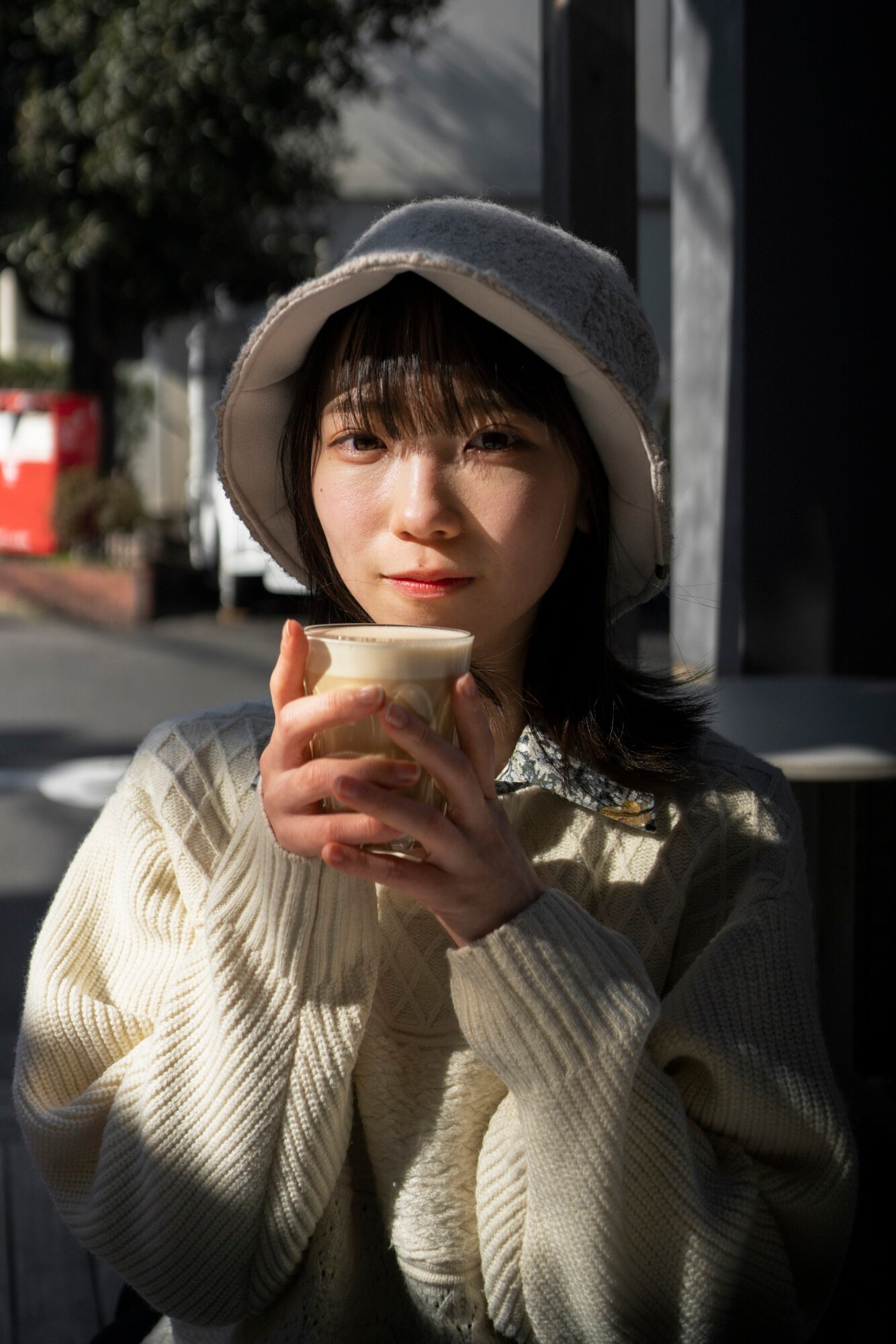 Person wearing a cream-colored knit sweater and a soft bucket hat, holding a cup with both hands close to their face as if enjoying a warm drink. The scene is outdoors in natural daylight, with a background of urban elements including a sidewalk, trees, and buildings. The overall mood is calm and cozy, suggesting a quiet moment of relaxation.