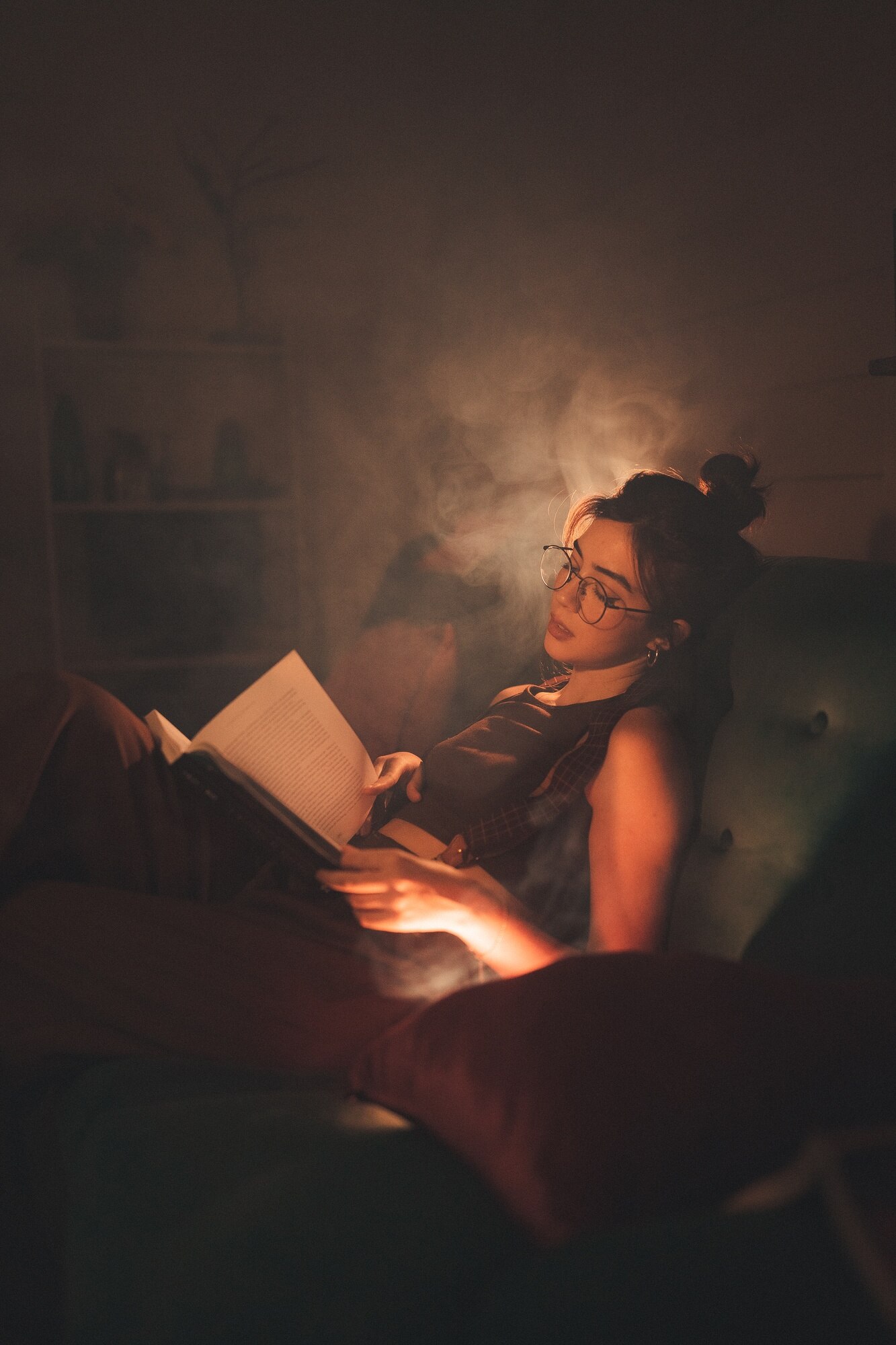 Woman reclining on a green sofa in a dimly lit room, holding an open book in her lap. Warm, soft light highlights her relaxed posture and creates a cozy, tranquil atmosphere. Wisps of smoke or mist drift around her, adding to the sense of calm and introspection. The background is softly blurred, with shelves and plants faintly visible, enhancing the intimate and peaceful mood.