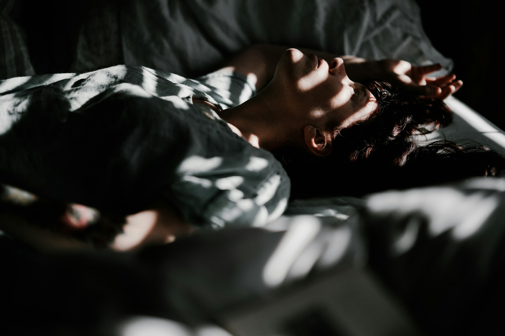 Person lying on a bed with dappled sunlight and shadow patterns across their face and body. The person appears to be resting or lost in thought, with one arm raised above their head. The room is dimly lit, creating a contemplative and tranquil atmosphere.