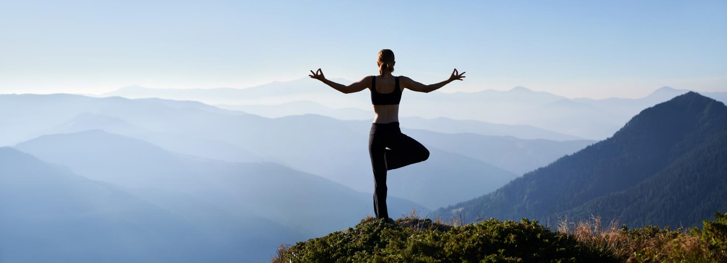 Person practicing yoga on a mountaintop at sunrise, standing in tree pose with arms extended and fingers forming mudras. The background features layers of misty blue mountains under a clear sky, creating a peaceful and uplifting atmosphere that emphasizes connection with nature and tranquility.