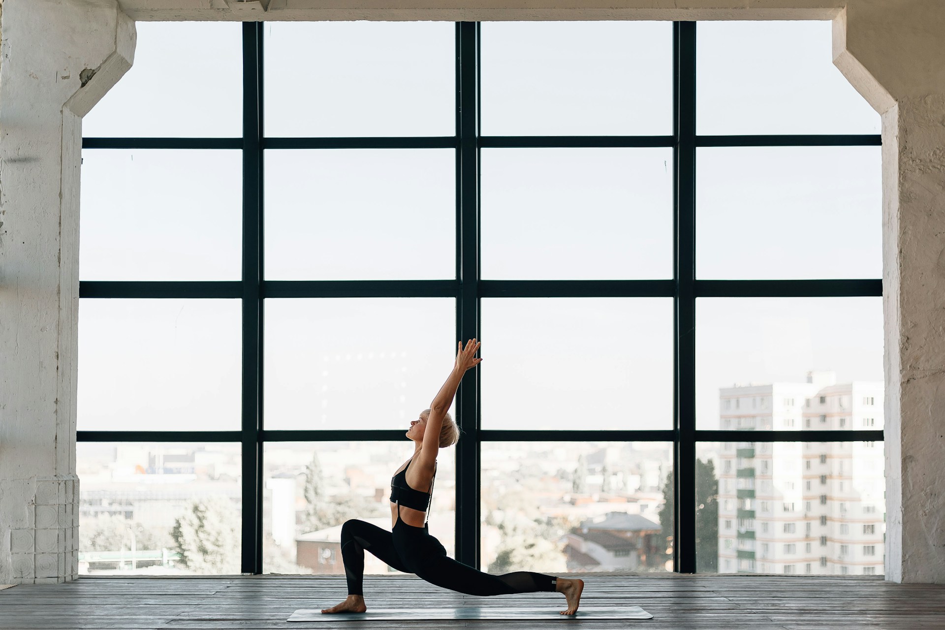 A woman in athletic clothing practices a yoga pose on a mat in a spacious room with large floor-to-ceiling windows. She is in a deep lunge with her arms extended overhead, looking upward. The bright, airy environment features a cityscape visible through the windows, creating a calm and focused atmosphere.