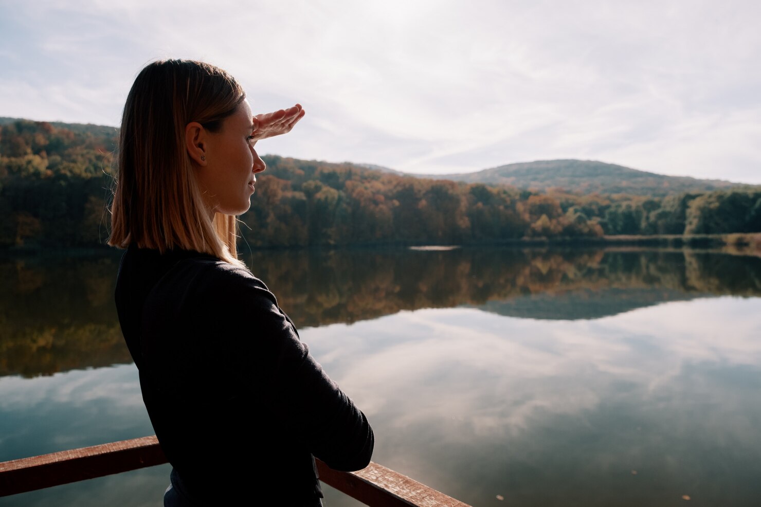 Woman with shoulder-length hair standing on a wooden railing overlooking a calm lake, shielding her eyes with her hand as she gazes toward distant forested hills under a partly cloudy sky. The scene is tranquil and contemplative, with reflections of the trees and sky mirrored on the water's surface, evoking a peaceful and thoughtful mood.