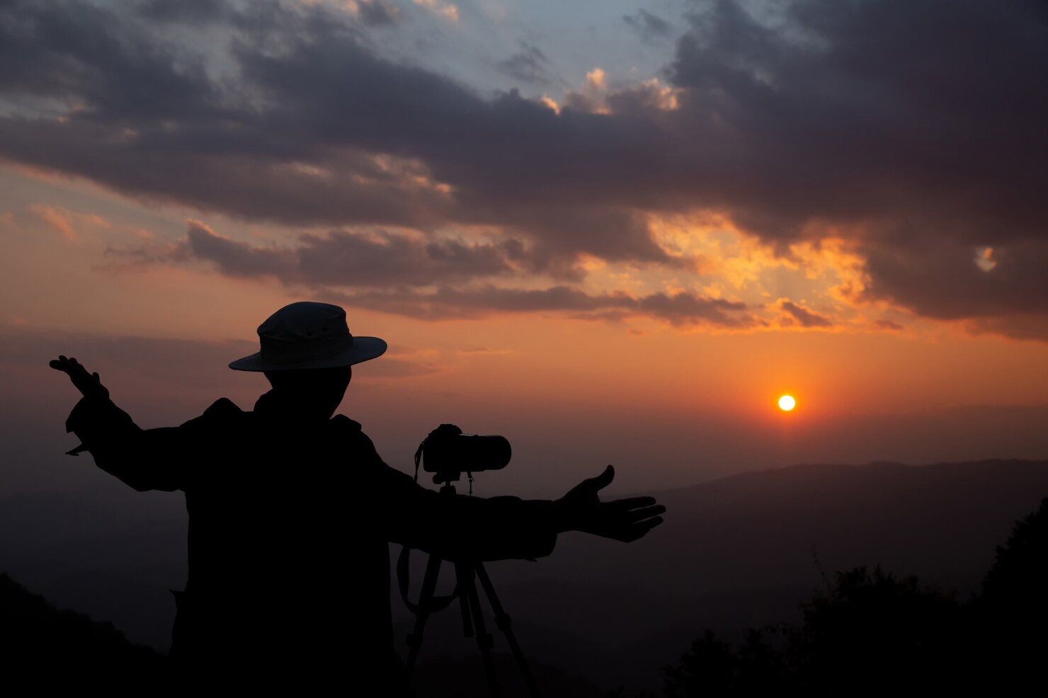 Silhouette of a person wearing a wide-brimmed hat standing next to a camera on a tripod at sunset, arms outstretched as if gesturing or presenting the view. The sky is filled with dramatic clouds and warm orange light as the sun sets over distant mountains, creating a peaceful and contemplative atmosphere.