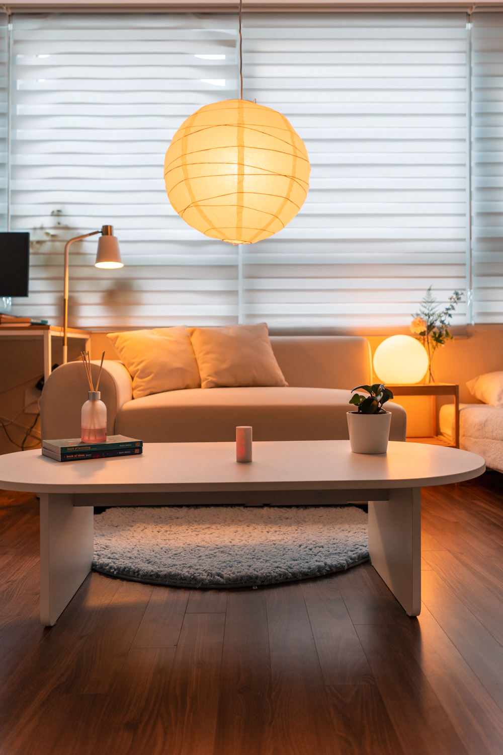 A minimalist living space featuring a white sofa with plush pillows, a low white coffee table holding books, a reed diffuser, and a small potted plant. A neatly made bed with light bedding sits nearby, and the room is softly illuminated by a large paper lantern and natural light filtering through white blinds. The environment feels calm, cozy, and organized, evoking a sense of peaceful relaxation.