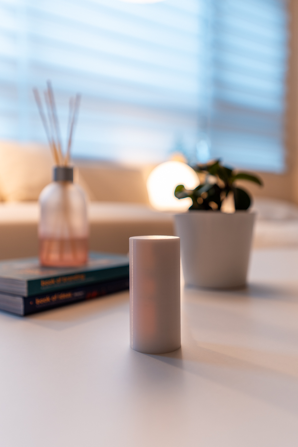 Lumaneos Light Controller. A white cylindrical object stands on a minimalist white table in a softly lit modern living room. Nearby are a small potted plant, a stack of books, and a translucent reed diffuser. The background features a sofa, large windows with blinds, and warm ambient lighting, creating a calm and cozy atmosphere.