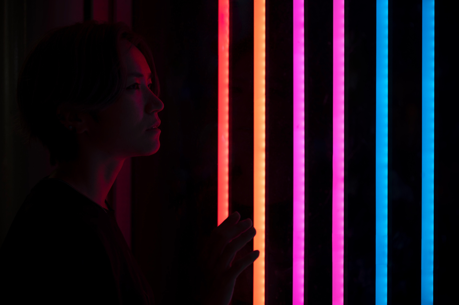 A person stands in a dark room facing a wall with vertical neon light strips in orange, pink, and blue. The person raises one hand toward the glowing lights, which cast colorful reflections on the surroundings. The environment feels moody and futuristic, evoking curiosity and contemplation.