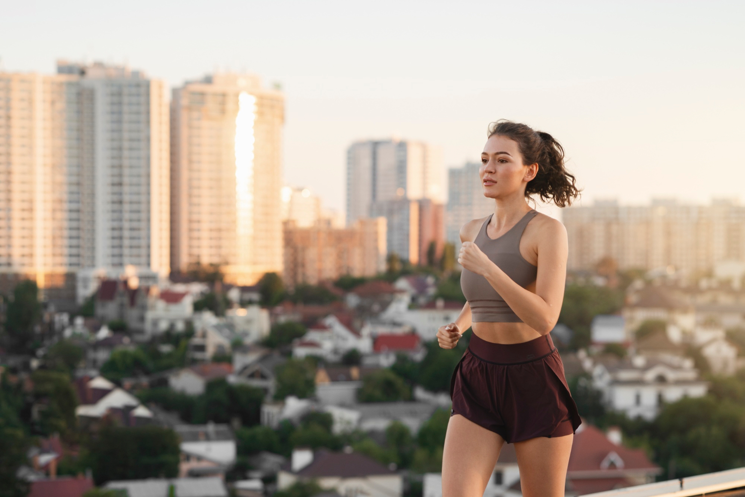 Woman in athletic wear jogging outdoors on a rooftop or terrace with a cityscape of tall apartment buildings and houses in the background. The scene is bathed in warm, soft sunlight, suggesting early morning or late afternoon. The overall mood is energetic and focused, with an urban environment providing a sense of openness and activity.