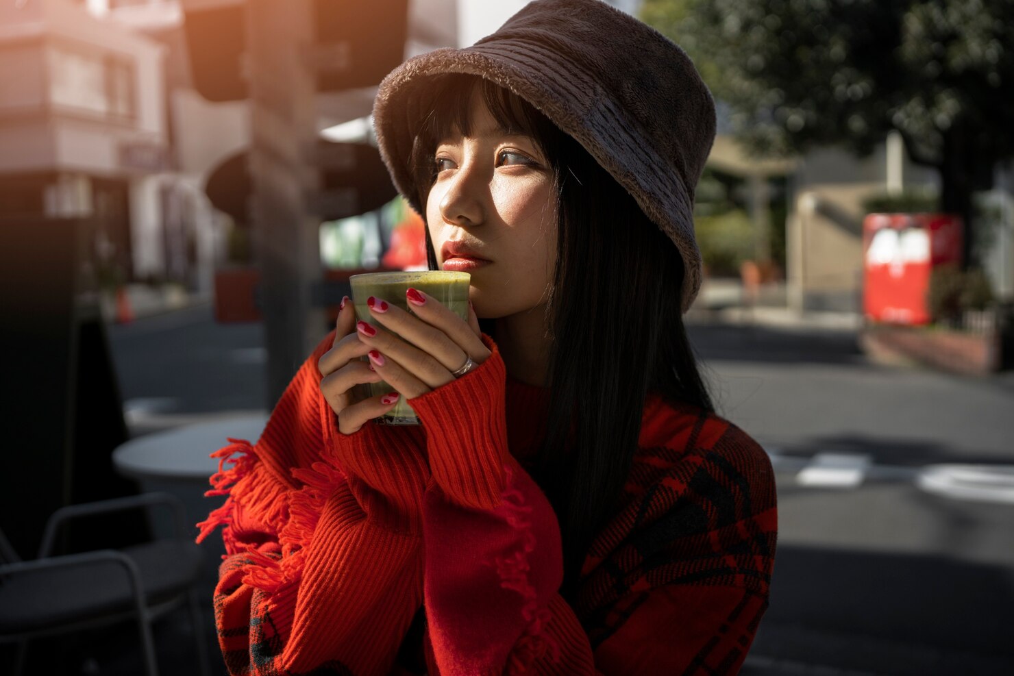 Person wearing a soft bucket hat and a bright red sweater with fringe details, holding a cup with both hands close to their face as if enjoying a warm drink. The scene is outdoors on a city street in natural daylight, with urban elements such as a sidewalk, trees, and buildings in the background. The overall mood is calm and cozy, suggesting a quiet moment of relaxation.