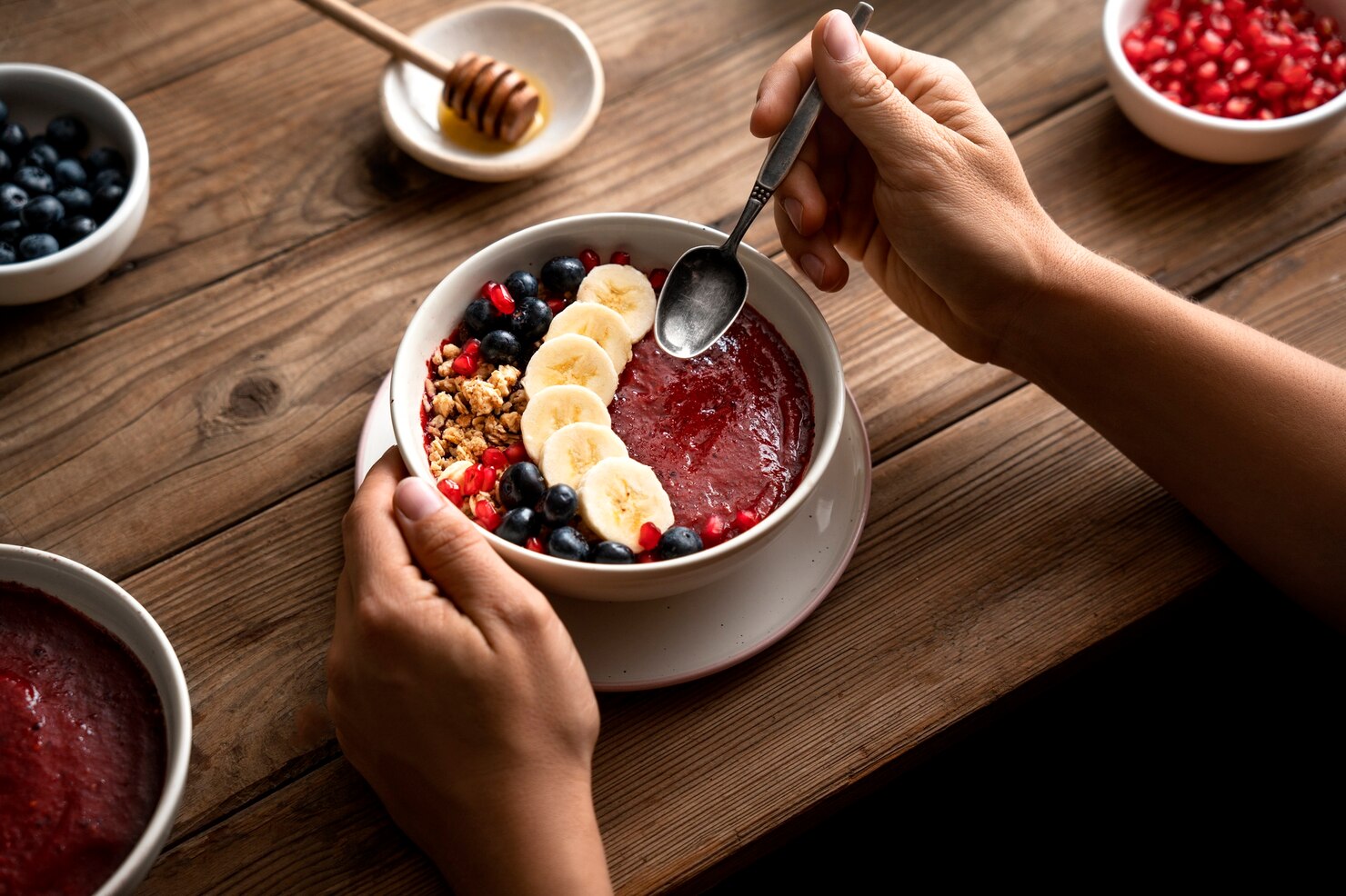 Hands holding a bowl of acai smoothie topped with banana slices, blueberries, granola, and pomegranate seeds on a rustic wooden table. A spoon is poised above the bowl, ready to eat. Surrounding the main bowl are small dishes with blueberries, pomegranate seeds, and honey, creating a cozy and inviting breakfast scene. The overall mood is warm and nourishing.