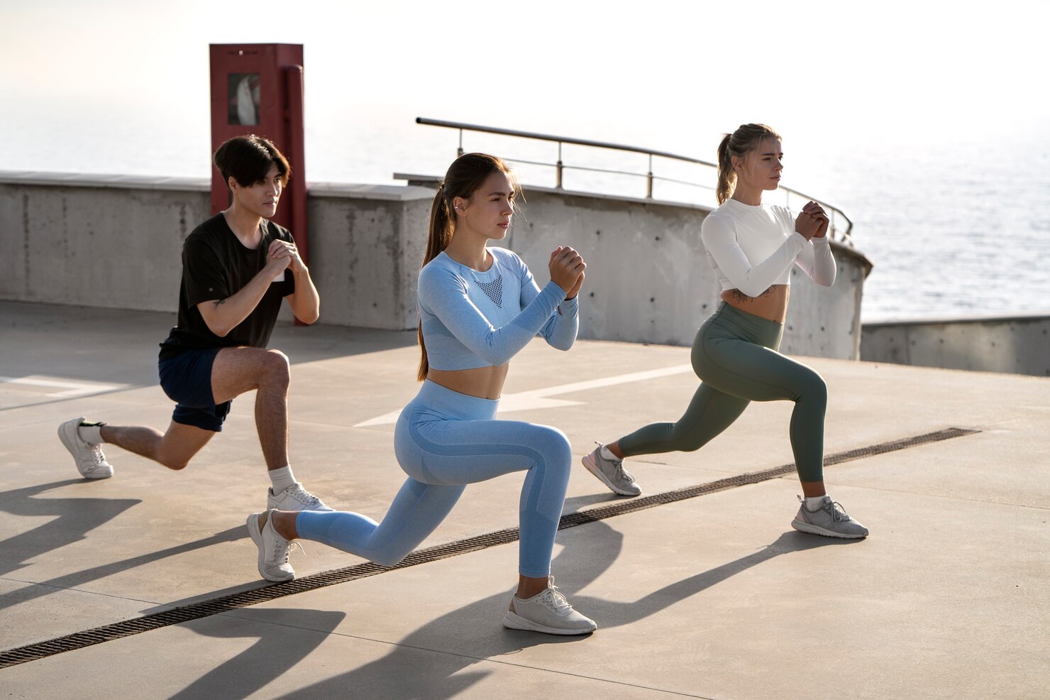 Three people in athletic wear performing forward lunges on a concrete surface outdoors near the ocean. They are positioned in a row, each with one knee bent and hands clasped in front of their chest, appearing focused and engaged in a group workout. The background features a low concrete wall, metal railings, and bright natural light, creating an energetic and motivating atmosphere.