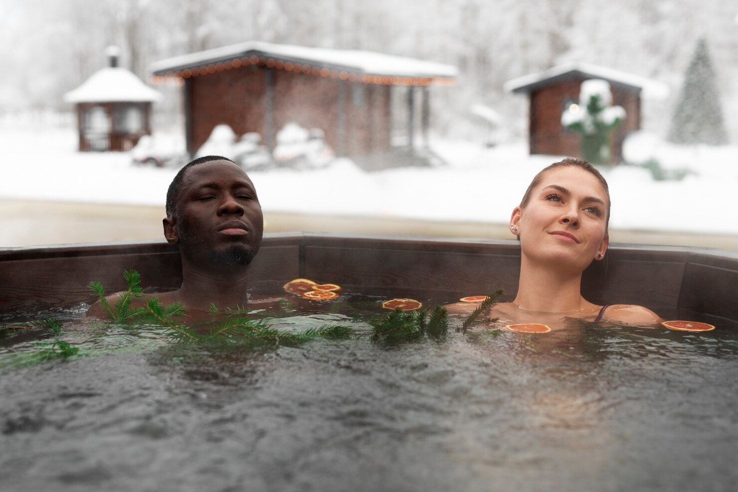 Two people relaxing in an outdoor hot tub surrounded by snowy scenery, with evergreen branches and slices of citrus fruit floating on the water. Wooden cabins and snow-covered trees are visible in the background, creating a cozy and tranquil winter atmosphere.
