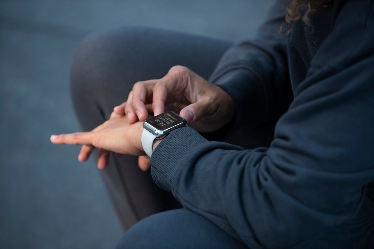 Person in dark athletic clothing sitting outdoors, interacting with a smartwatch on their wrist. The focus is on their hands and the smartwatch screen, which displays fitness tracking information. The background is blurred, suggesting an urban or park setting. The mood is attentive and engaged, emphasizing health monitoring and technology use.