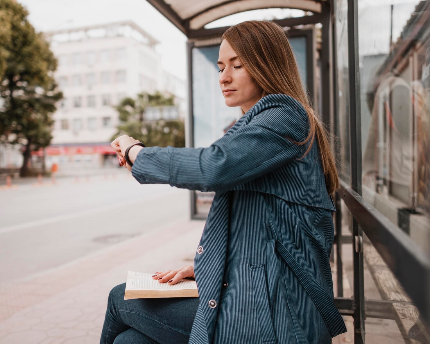 A college-age commuter checks the time while waiting at a bus stop with a book on her lap.
