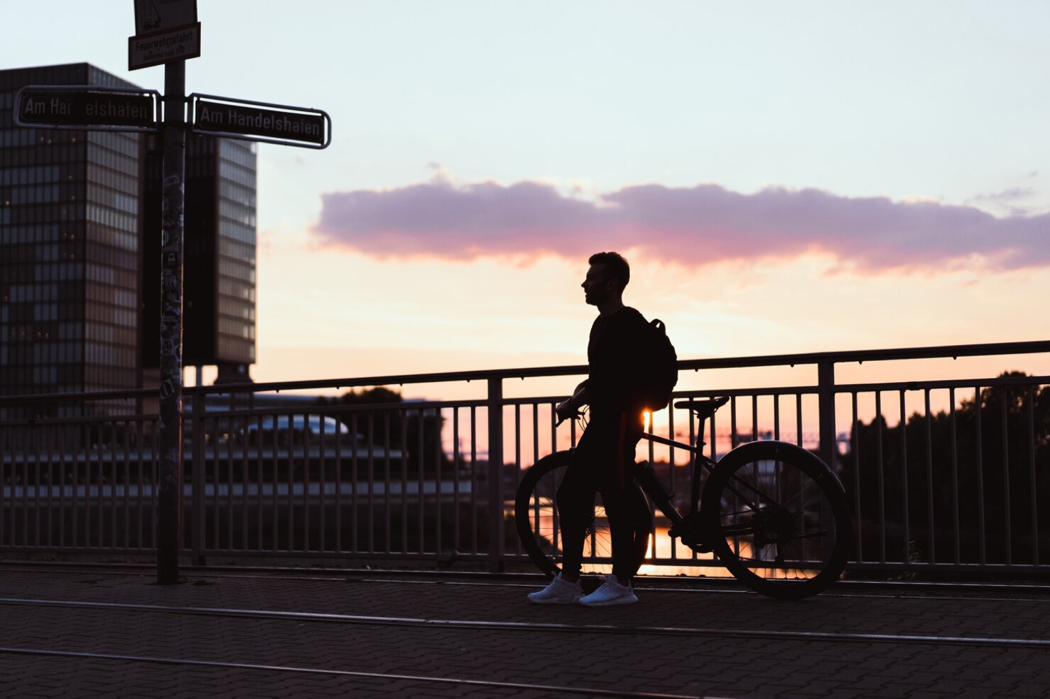 A commuter with a backpack walks a bicycle across a city bridge in warm dawn light.