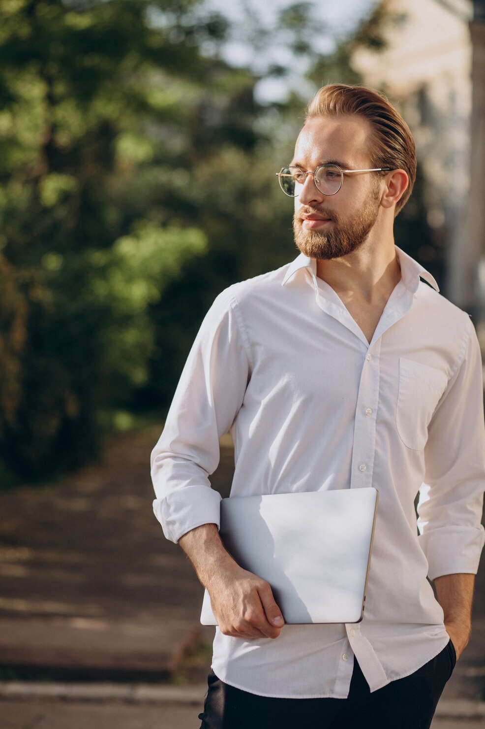 A young professional carries his laptop while walking toward work in warm morning light.