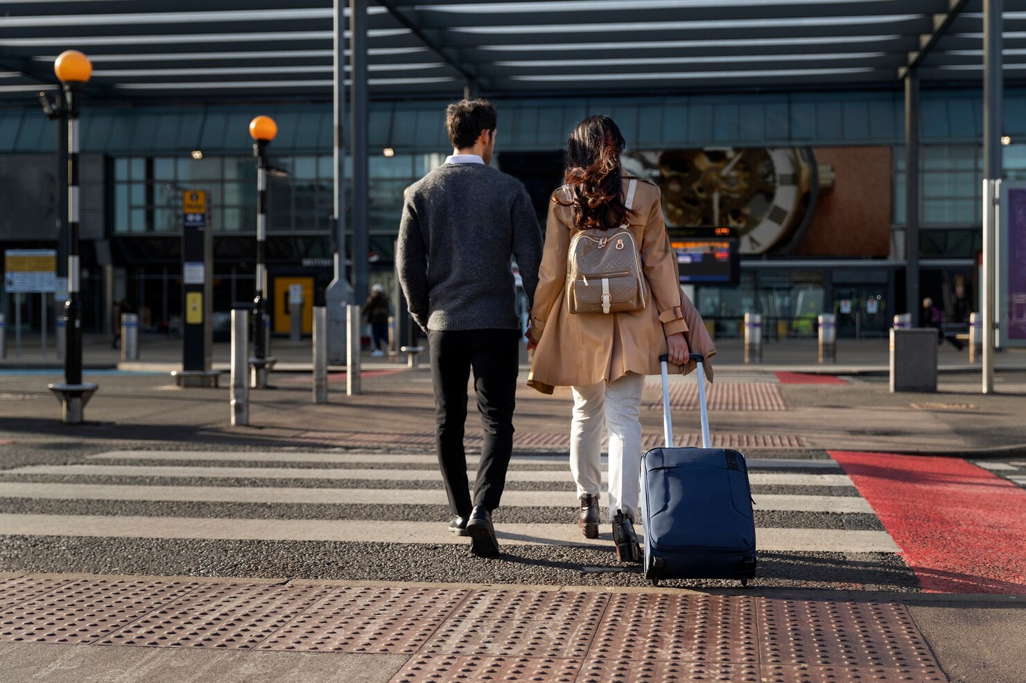 Man and woman walking side by side toward an airport entrance, seen from behind. The woman is pulling a blue rolling suitcase and wearing a tan coat with a light backpack, while the man is dressed in dark pants and a gray sweater. They are crossing a zebra-striped crosswalk with tactile paving under a large modern canopy. The environment is bright and orderly, with signs, bollards, and glass walls visible, conveying a sense of anticipation and travel.