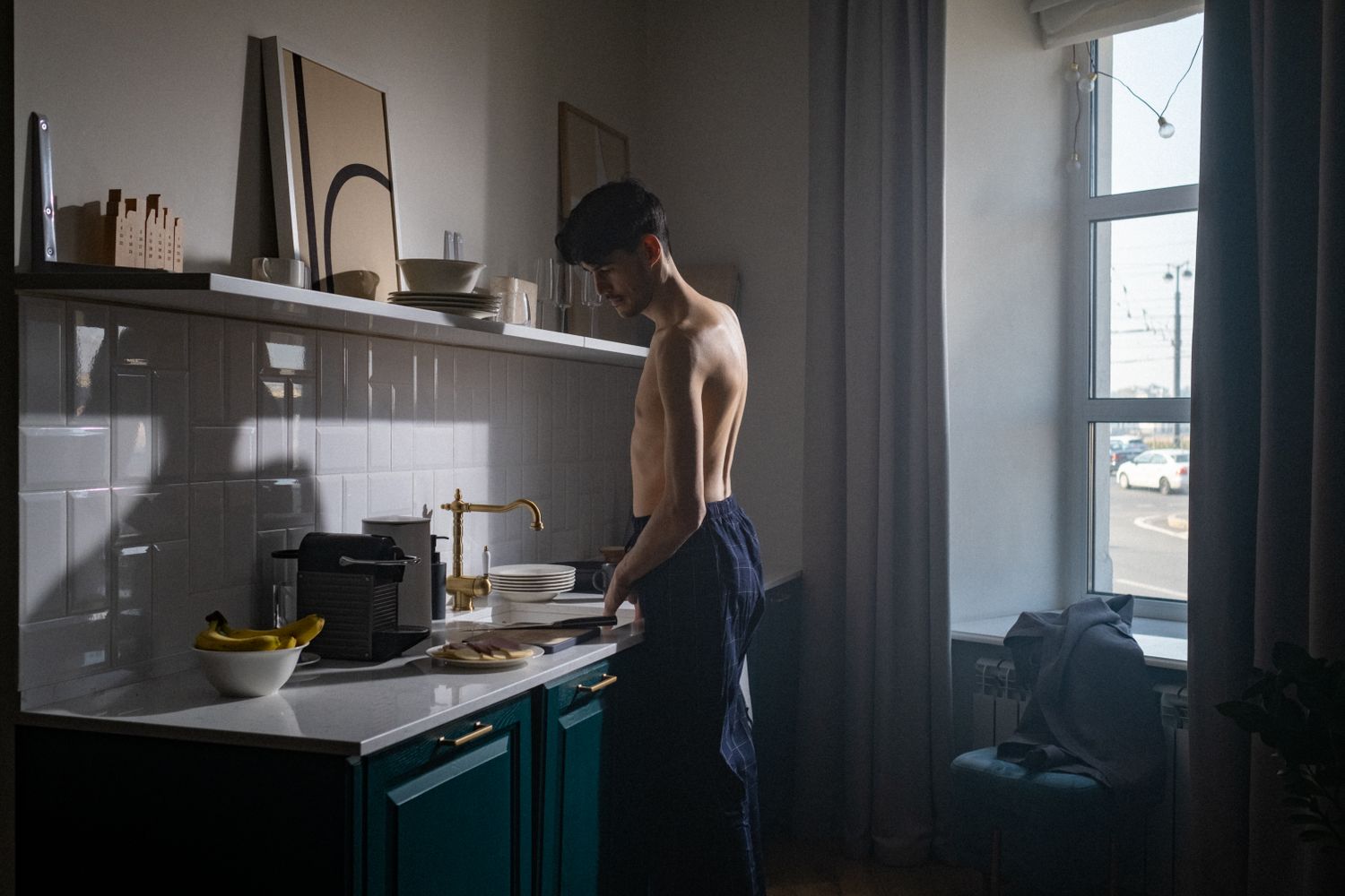 A man prepares breakfast in a dim kitchen as morning light enters through the window.