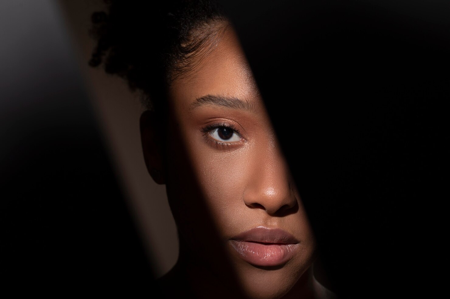 Person with dark curly hair sits in a dimly lit room, with soft shadows and a warm, subdued atmosphere. The background is mostly dark, creating a sense of privacy and introspection.