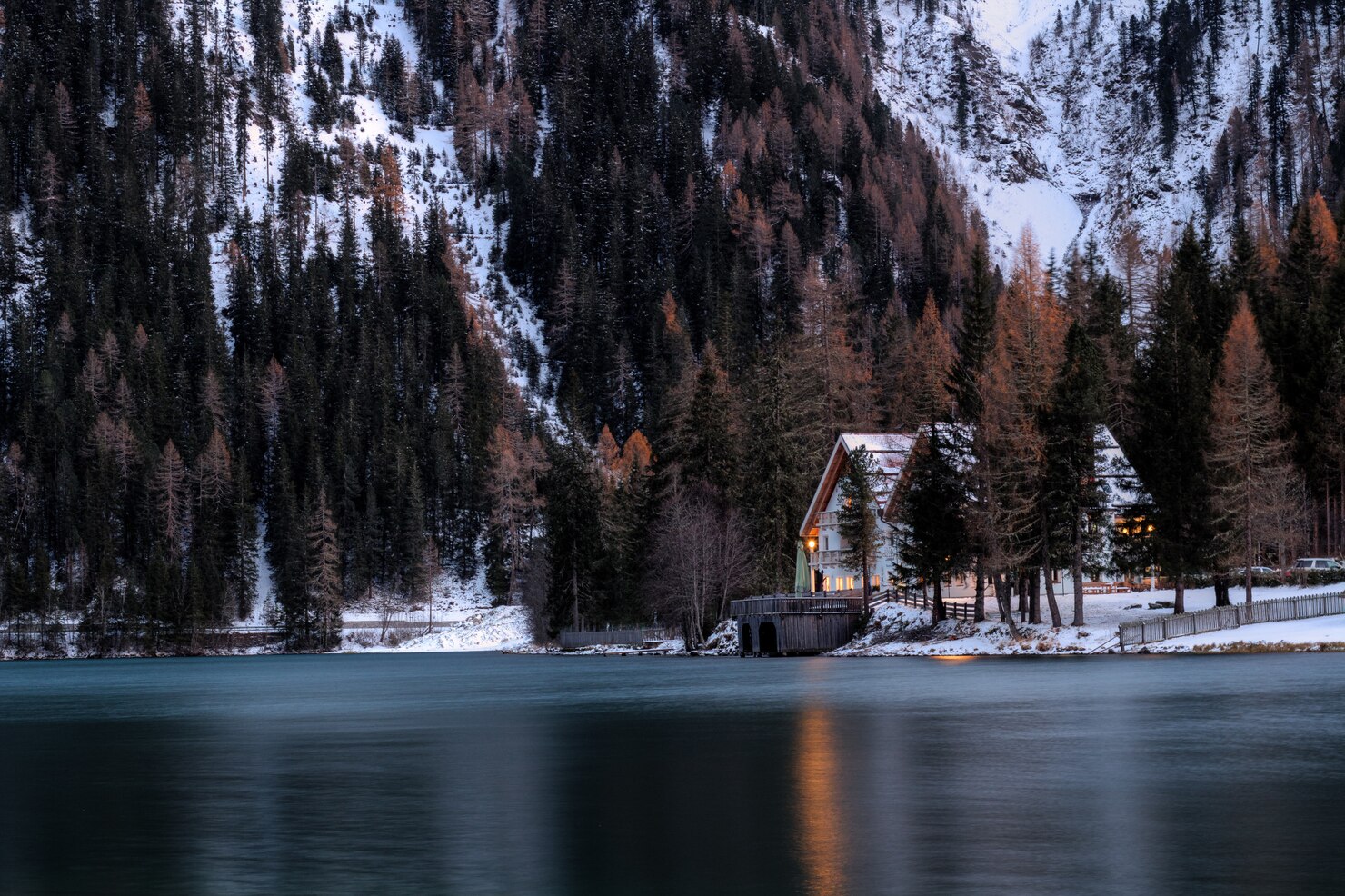 Snow-covered mountain landscape with dense evergreen forest surrounding a calm lake. A wooden cabin with warm lights glowing from the windows sits at the water's edge, partially hidden by tall trees. The scene is tranquil and serene, with reflections of the cabin and trees on the still surface of the lake, evoking a peaceful and cozy winter atmosphere.