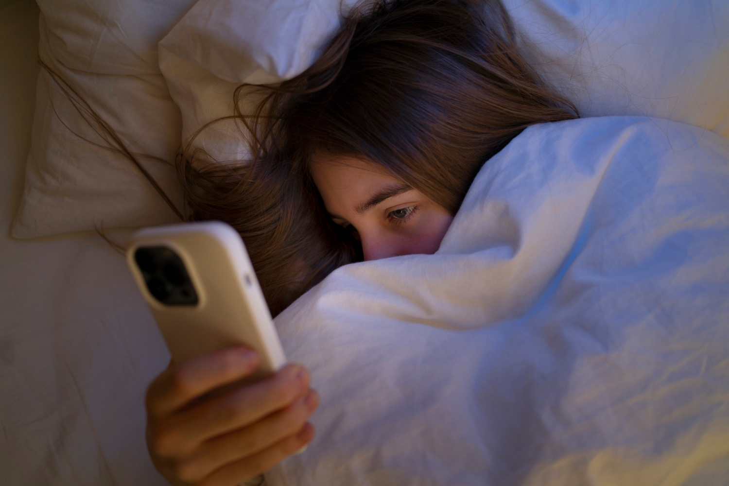 A person lies awake in bed at night looking at a phone while wrapped in white bedding.