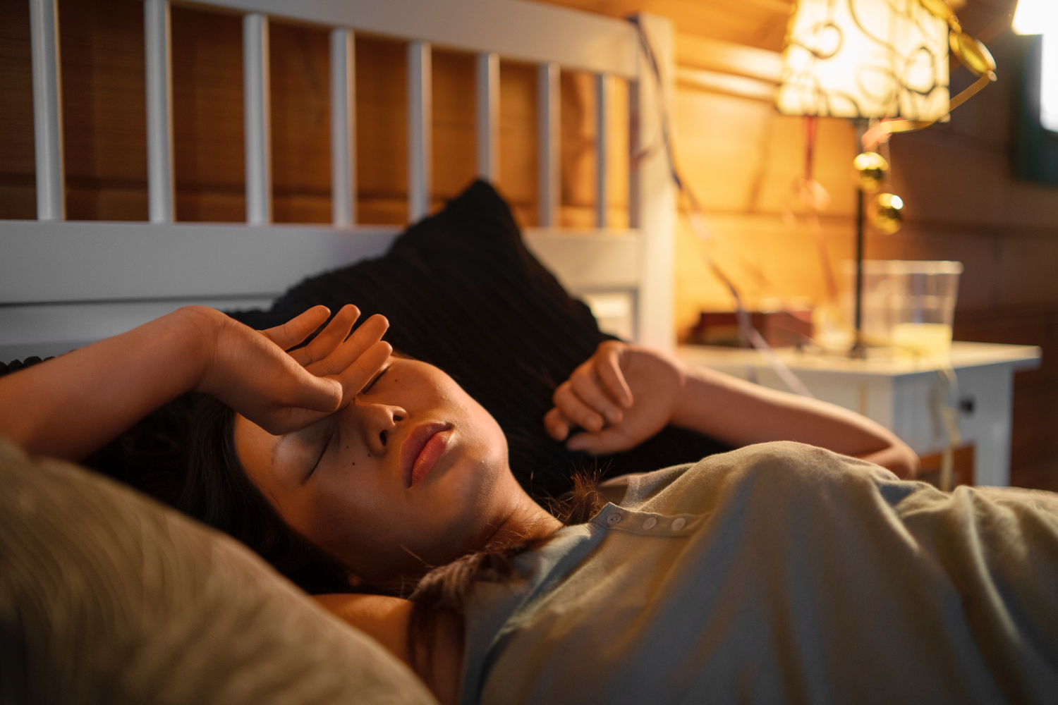 A person lies on a bed in a warmly lit wooden bedroom, wearing a light gray shirt and resting one arm across their forehead. The bed has white sheets and dark pillows, and a nightstand beside the bed holds a lamp with a decorative shade and a glass of water. The environment feels cozy and relaxed, suggesting rest or winding down at the end of the day.
