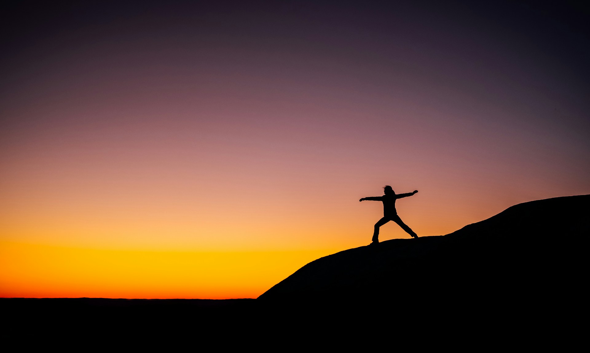 A person stands in a wide-legged pose on a rocky hilltop at sunset, arms extended outward. The sky transitions from deep purple at the top to vibrant orange near the horizon, creating a dramatic and peaceful atmosphere. The silhouette is sharply defined against the colorful sky, evoking a sense of freedom, balance, and tranquility.
