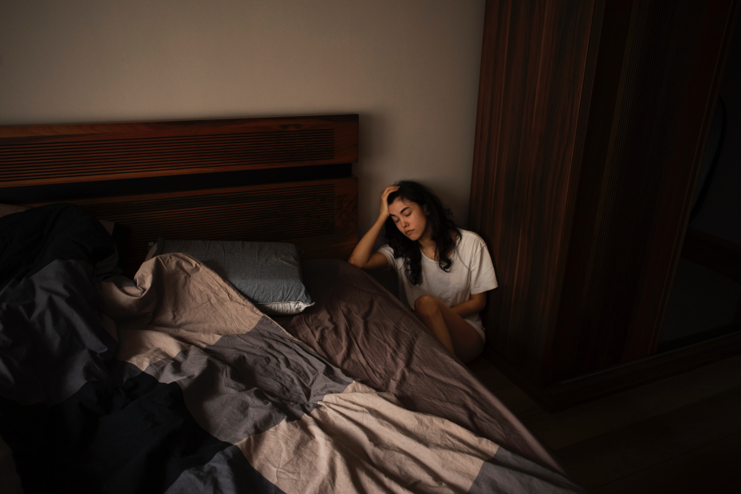 A person with long dark hair sits on the floor next to a bed in a dimly lit bedroom, wearing a white t-shirt and resting their head in one hand. The bed has rumpled sheets and pillows, and a wooden headboard is visible. The room features wooden furniture and soft lighting, creating a quiet and somber atmosphere that suggests fatigue or contemplation.