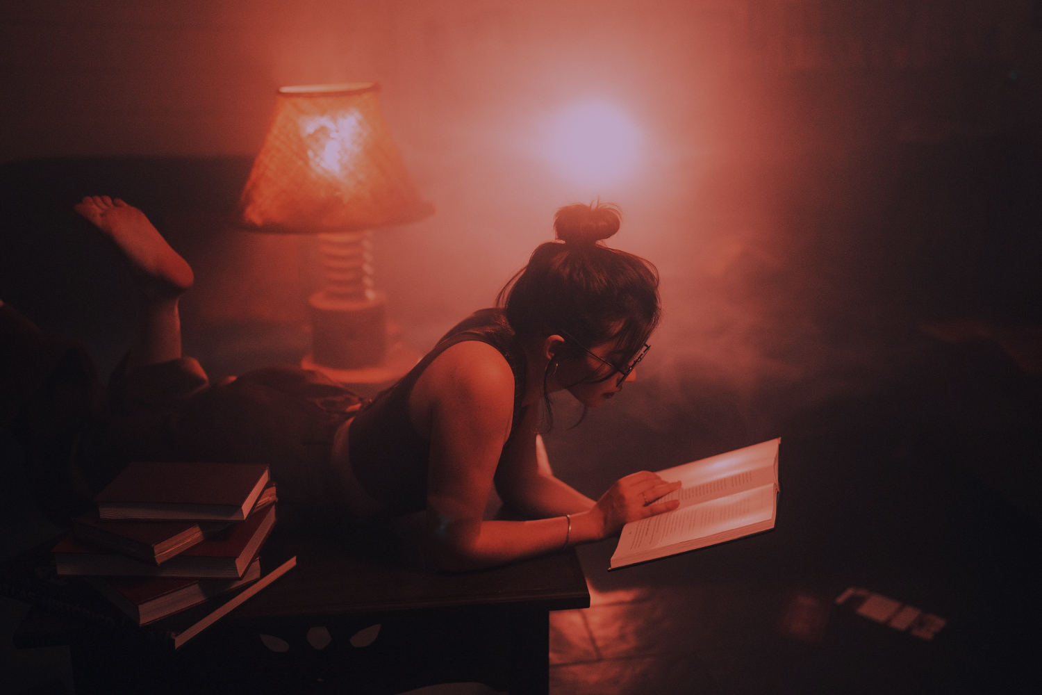 A woman lies on her stomach on a wooden table, reading an open book in a dimly lit room. She is surrounded by several stacked books, and a warm lamp glows nearby, casting soft light and creating a cozy, tranquil atmosphere. The scene feels calm and introspective, suggesting quiet study or relaxation.