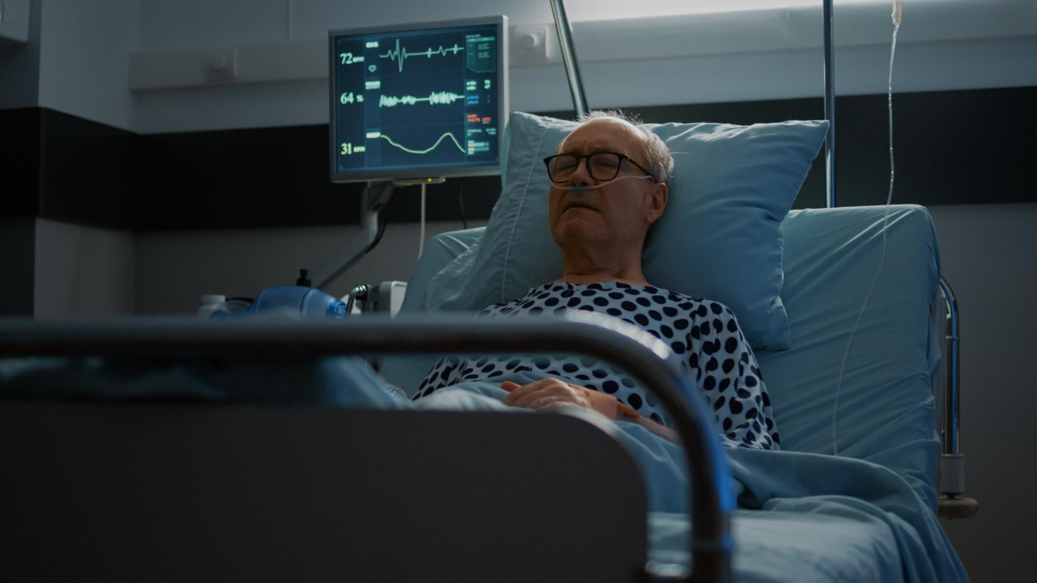 A person lies in a hospital bed wearing a patterned hospital gown, hands resting on their lap. Medical monitoring equipment beside the bed displays vital signs including heart rate, oxygen saturation, and pulse, with green and yellow lines on a dark screen. The room is dimly lit with clinical lighting, creating a serious and somber atmosphere that suggests a medical situation or recovery.