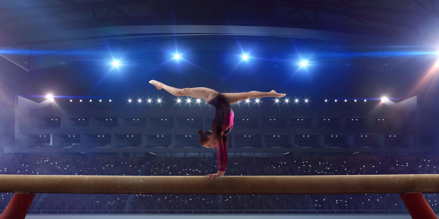 A gymnast in a sparkling maroon leotard performs a handstand split on a balance beam in a large indoor arena. The gymnast balances with arms straight and legs extended in a perfect split, demonstrating strength and flexibility. Bright stage lights illuminate the scene, and a large audience fills the stadium seats in the background, many holding up glowing phones. The atmosphere is dramatic and focused, capturing the intensity and skill of a gymnastics competition.