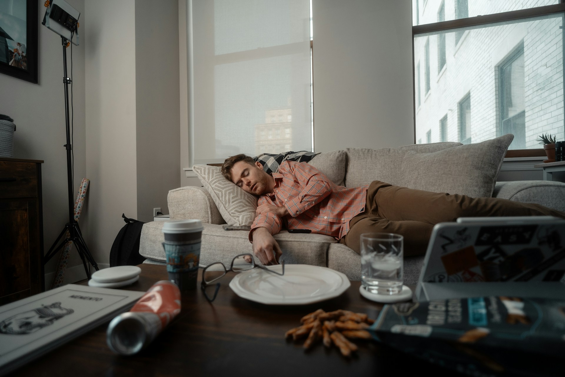 A person wearing a pink plaid shirt and brown pants lies on a beige sofa in a modern living room, appearing relaxed or tired. The coffee table in the foreground holds a plate with snacks, a glass of water, a coffee cup, a pair of eyeglasses, and a laptop with stickers. Large windows let in natural light, and the overall atmosphere feels casual and slightly cluttered, suggesting a break or downtime at home.