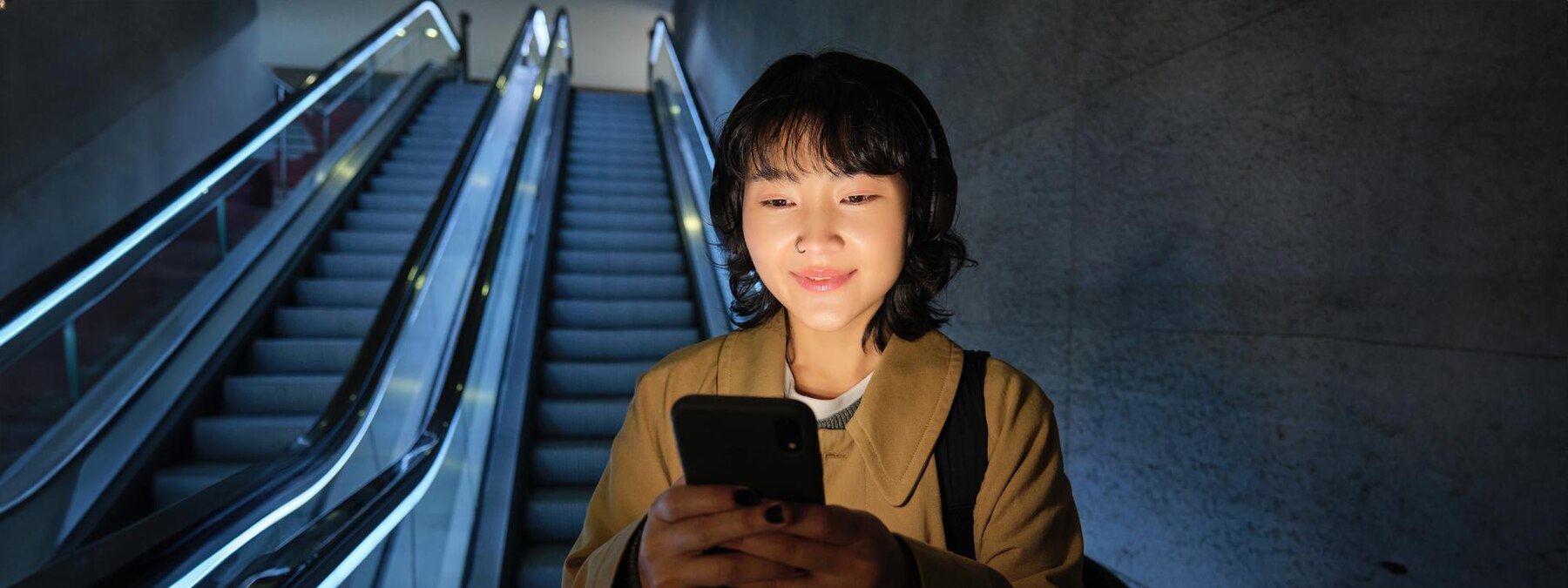 A person stands at the base of an escalator in a dimly lit modern indoor space, holding a smartphone and wearing headphones. The escalator is illuminated with blue-tinted lights and leads upward into the background. The environment feels quiet and slightly moody, suggesting a moment of focus or waiting in a transit area or public building.