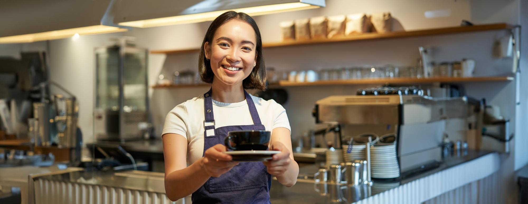 A barista wearing a white t-shirt and dark apron stands behind a counter in a modern coffee shop, holding out a black cup and saucer with both hands as if serving a customer. The background features a professional espresso machine, stacks of cups, glassware, and shelves with coffee supplies. The environment is clean, organized, and warmly lit, creating a welcoming and professional atmosphere.