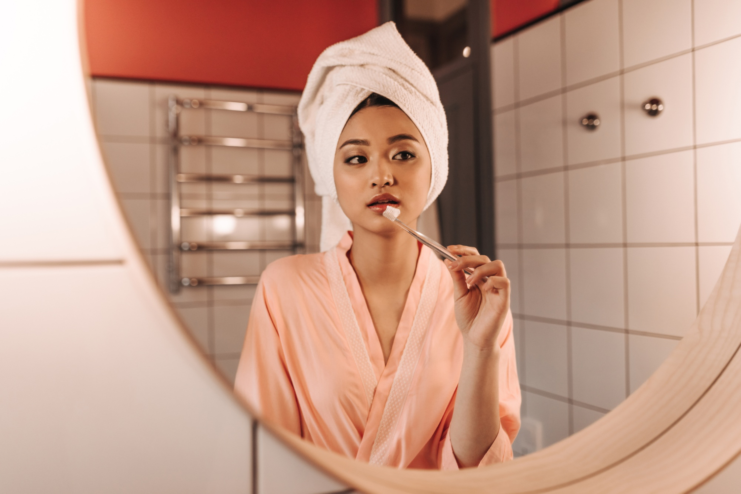 A woman in a robe brushes her teeth in front of a brightly lit bathroom mirror at night.