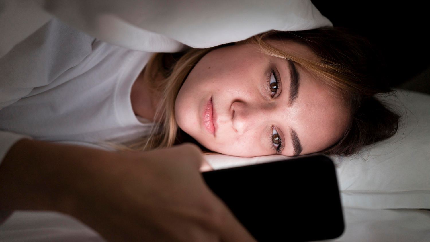 A close-up of a woman lying in bed at night looking at a glowing smartphone under the covers.