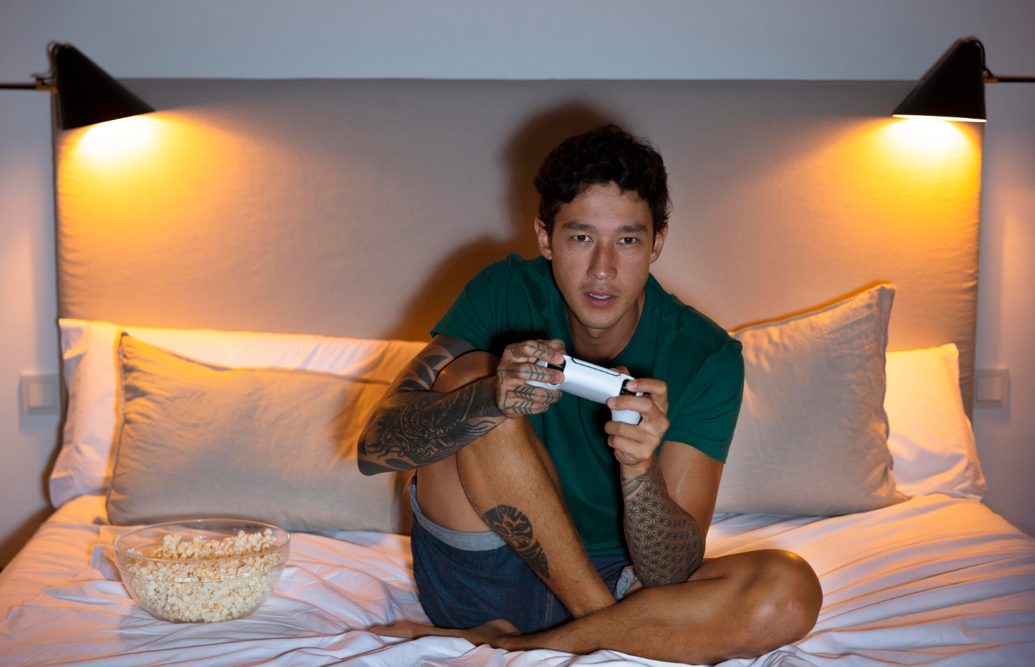 A man sits on a bed at night holding a video game controller beside a bowl of popcorn under bright bedside lamps.