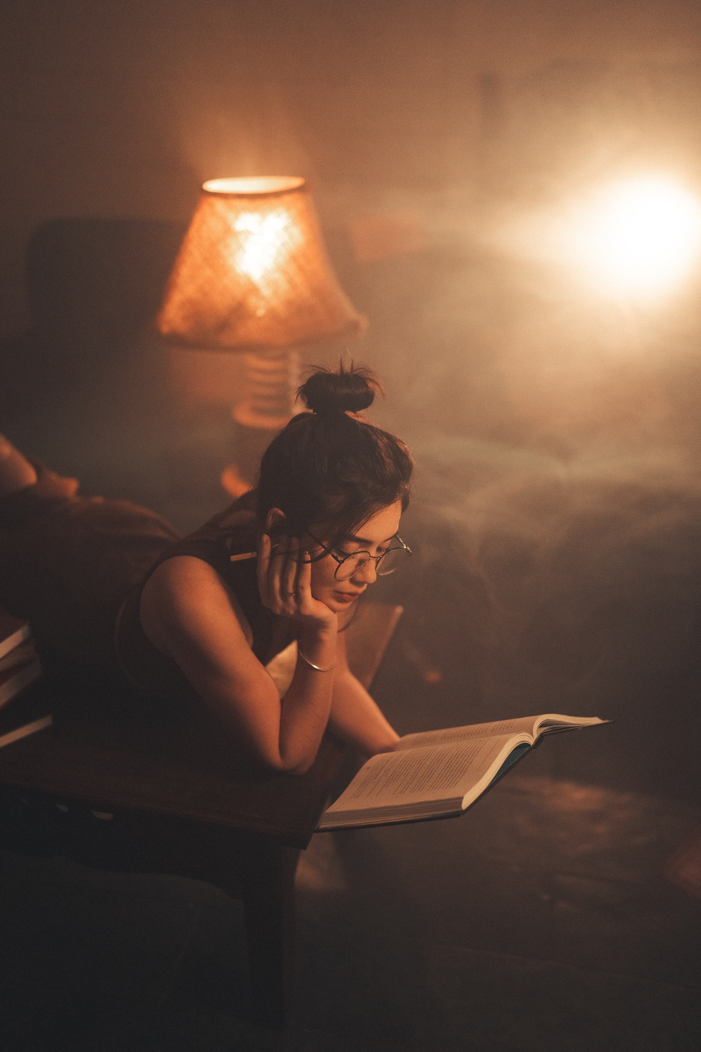 A woman leans over an open book in warm, dim lamplight, looking drowsy as she reads in a quiet room.