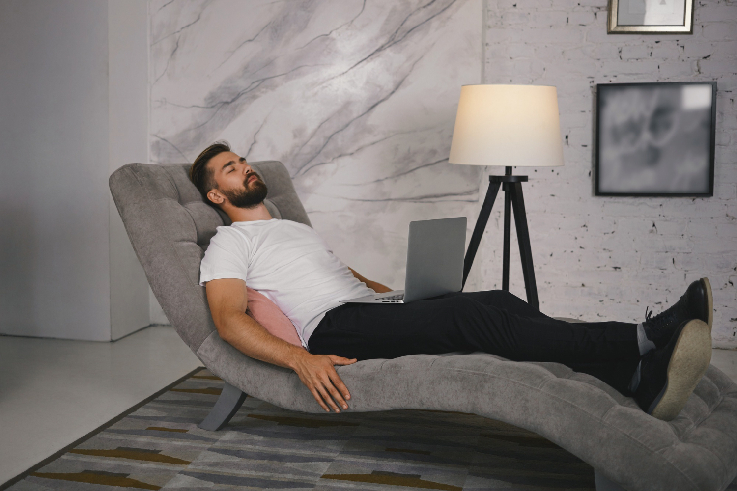 A man reclines on a modern gray chaise lounge with a laptop resting on his lap, appearing relaxed and possibly taking a break or resting. He wears a white t-shirt and black pants, with one arm resting on a pink pillow and the other on the lounge. The room features a marble accent wall, a floor lamp, framed art on a white brick wall, and a patterned rug, creating a calm and contemporary atmosphere.
