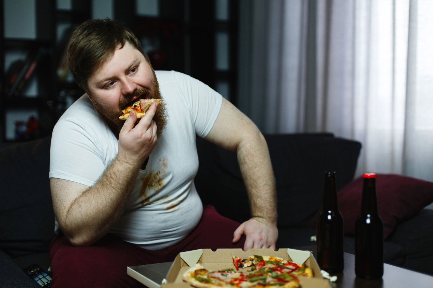 A person wearing a white t-shirt with food stains sits on a dark sofa, eating a slice of pizza from an open box on the coffee table. Two dark glass bottles are on the table next to the pizza. The room is dimly lit with shelves and curtains in the background, creating a casual and relaxed atmosphere that suggests a quiet night in.