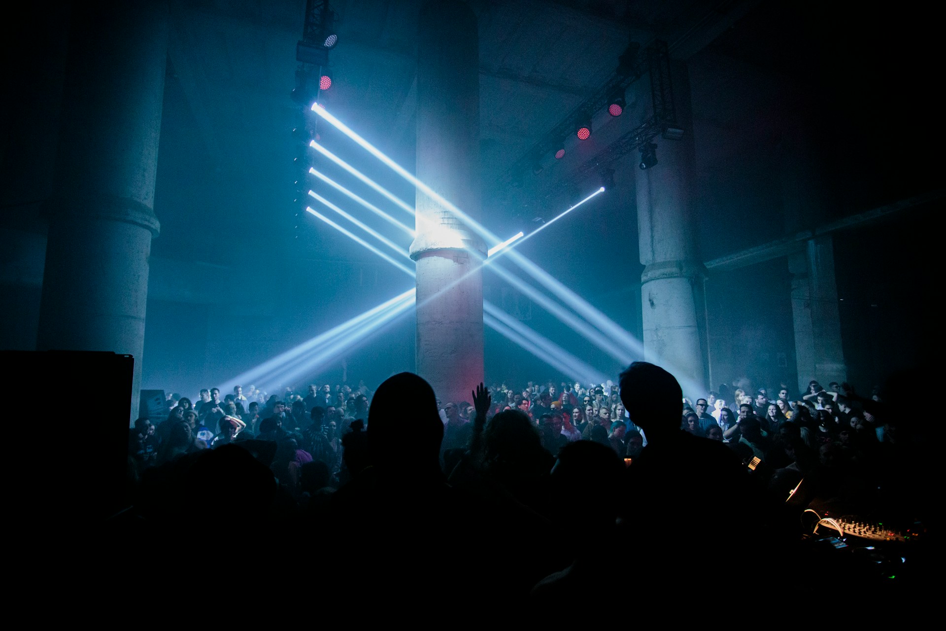 A large crowd of people gathers in a dark, industrial space with tall concrete pillars, illuminated by dramatic beams of white stage lights cutting through haze. The audience faces a DJ or performer at the front, with silhouettes visible in the foreground. The environment feels energetic and immersive, suggesting a lively music event or nightclub atmosphere.