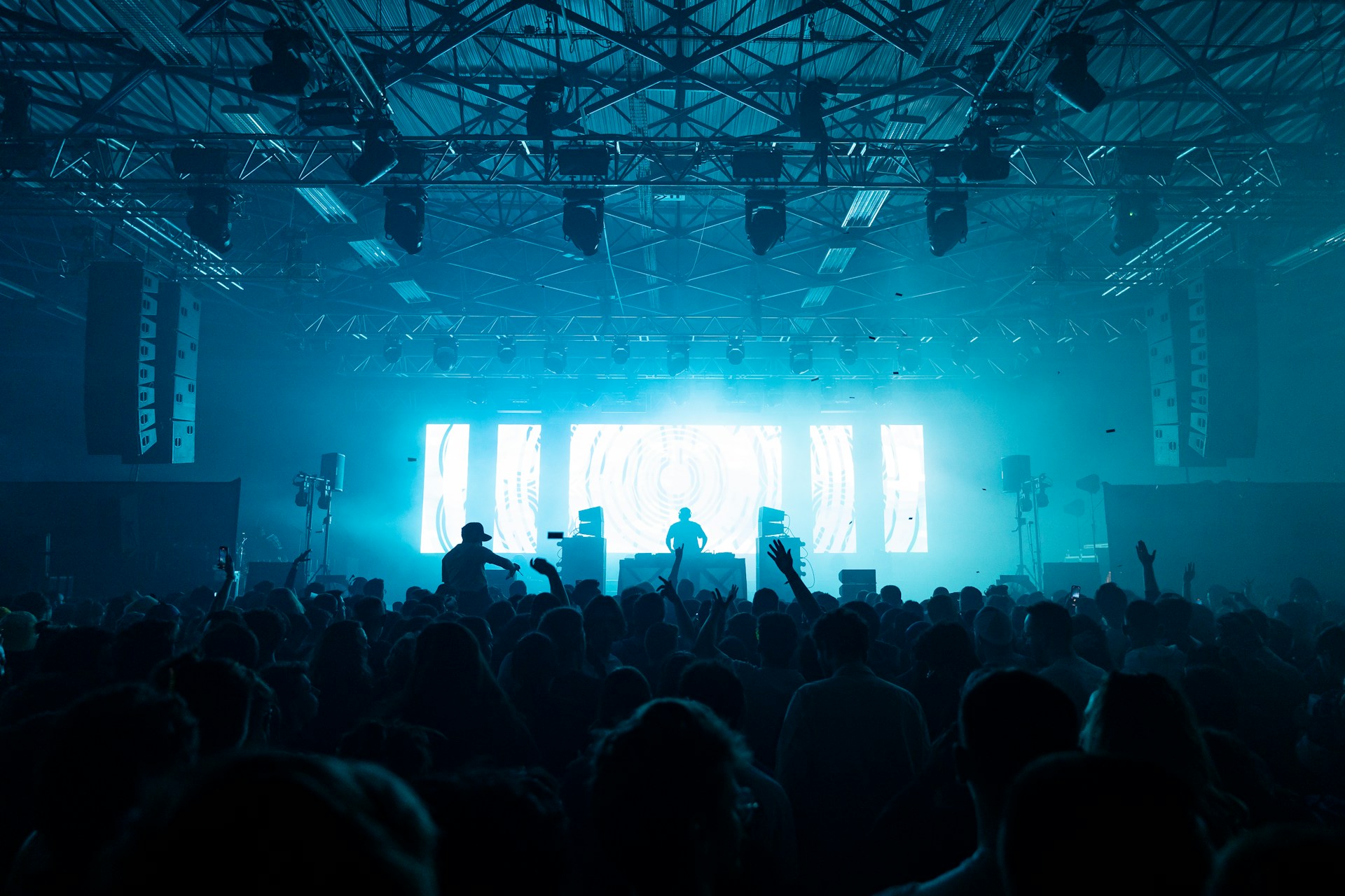 A DJ stands on stage in front of a large illuminated screen displaying abstract blue and white patterns, performing for a crowd of people in a dark indoor venue. Audience members raise their hands and move to the music, with silhouettes visible against the vibrant lights. The environment features an industrial ceiling with exposed beams and stage lighting, creating an energetic and immersive atmosphere that suggests excitement and celebration at a live music event.